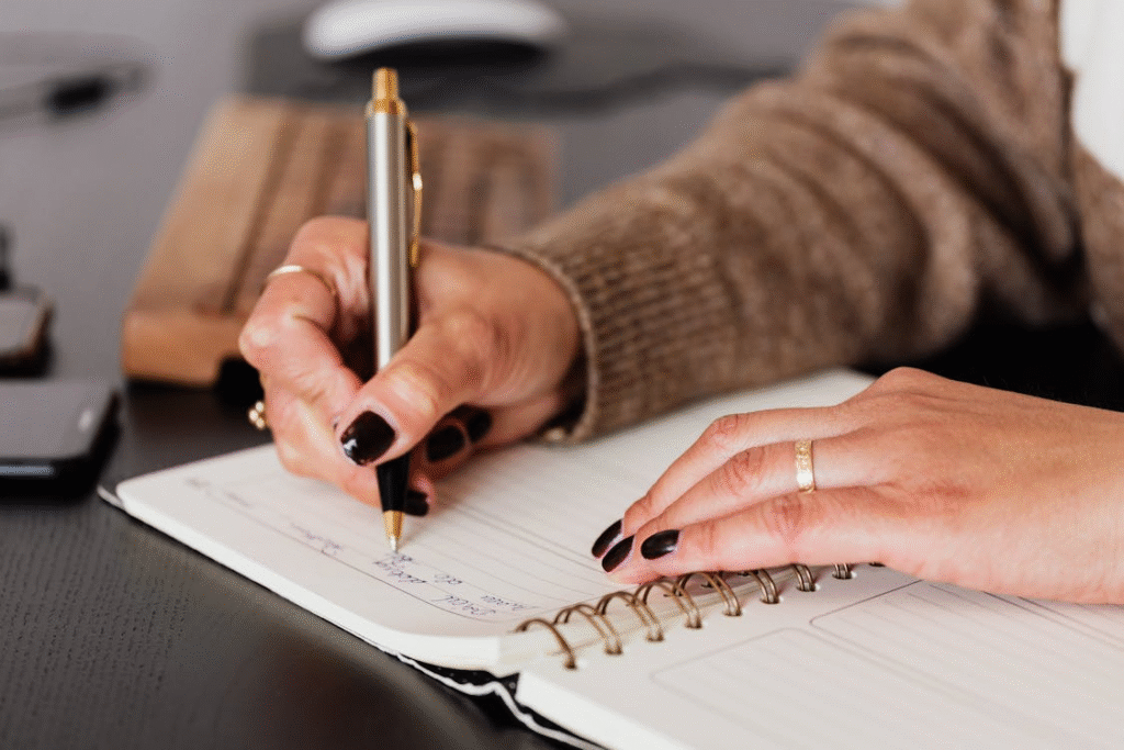 Close-up of woman writing in a spiral notebook at a desk.