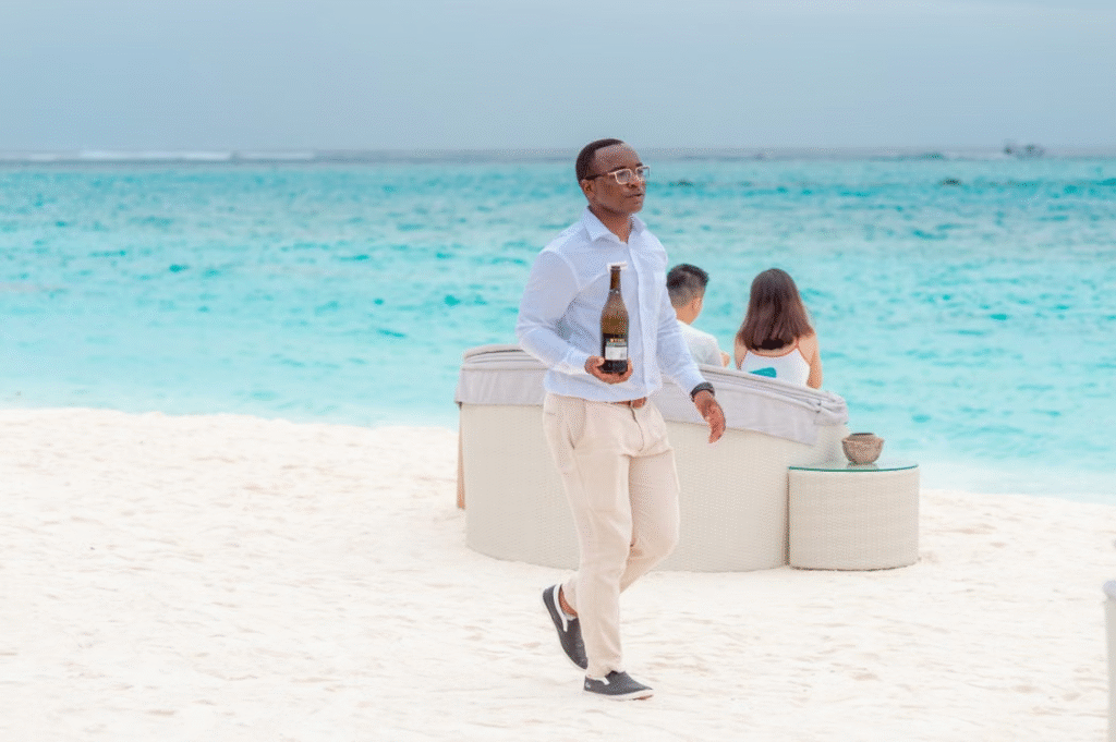 A waiter carrying a bottle on a sunny beach with people relaxing by the sea.