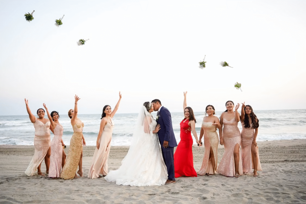Bride and groom kissing on a beach surrounded by joyful bridesmaids during sunset.